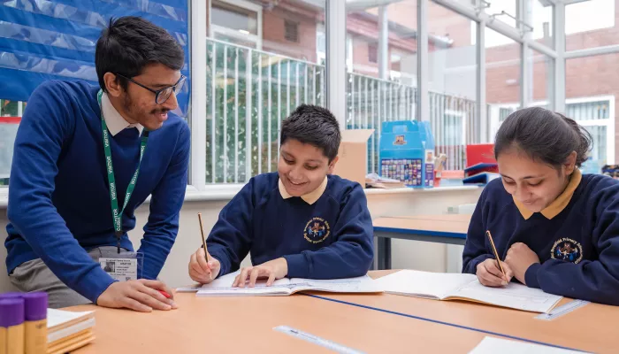 A male tutor with black hair and glasses, wearing a blue jumper, is leaning over a desk. Two primary aged pupils, a boy and a girl, wearing dark blue school jumpers are holding pencils and writing in exercise books. Everyone is smiling.
