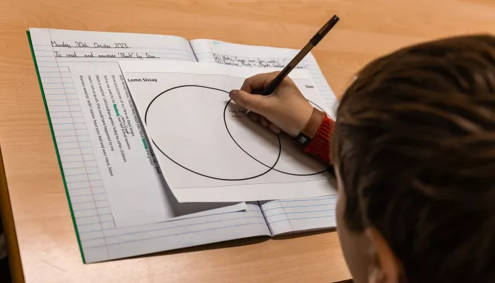 A close up photo of a school exercise book on a brown wooden desk. The exercise book is open to a Venn diagram and a male secondary student is using a pen to complete the Venn diagram. Only the back of his head is visible in the shot.