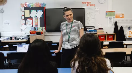 A young female tutor with dark hair, tied in a bun, wearing a striped t-shirt, is standing talking to two secondary-aged female pupils who are sat at a desk. The backs of the pupils' heads are facing the camera. Both pupils have long dark hair.