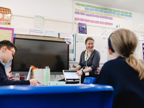 A female tutor is standing in front of a whiteboard, next to a large TV that is turned off. She is teaching two primary-aged pupils, whose faces are not visible. The tutor is smiling.