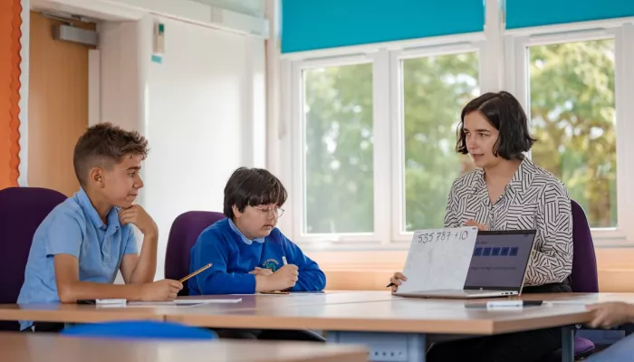 two primary school boys wearing blue jumpers, receiving tutoring from a female tutor with short dark hair