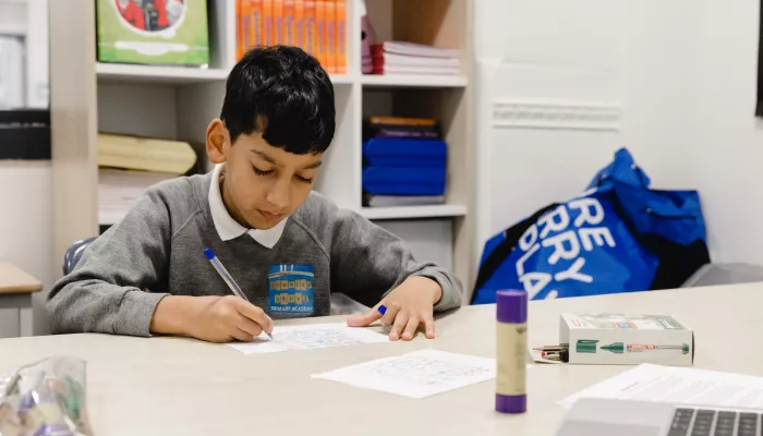 A primary school-aged boy wearing a grey school jumper is sat at a desk. He is writing with a pen on a sheet of paper and concentrating hard.