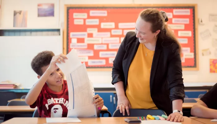 On the right of the image a female tutor wearing a yellow top and black blazer is standing at a desk. Sat next to her at the desk is a male pupil wearing a red t-shirt. He is holding up a large sheet of paper. The tutor and the pupil are both smiling.