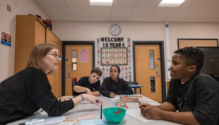 A female tutor wearing glasses is sat with three primary aged pupils around a desk. She is explaining a concept. The pupils are listening. In the back there is a colourful 'Welcome to Year 6' display on the wall.