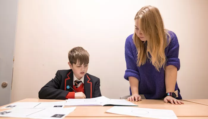 A secondary school-aged male pupil wearing a black blazer and red school jumper is sat at a desk. To his right is standing a female tutor with long blonde hair who is wearing a purple jumper. The boy is reading aloud to his tutor.