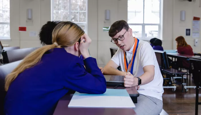 To the left of the picture two secondary school-aged female pupils wearing purple blazers are sat at a desk with their backs to the camera. On the other side of a desk a young male tutor wearing a white t-shirt and glasses is sat. He is looking down at a laptop and explaining a concept to the pupils.