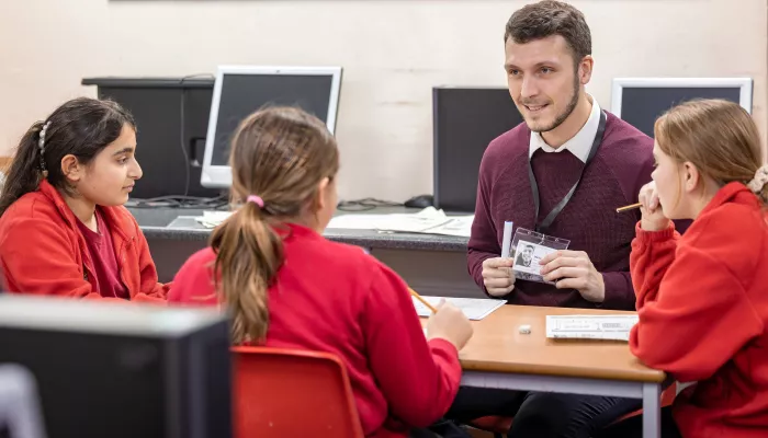 A male tutor wearing a burgundy jumper is sat tutoring a group of primary school-aged pupils wearing bright red school uniforms.