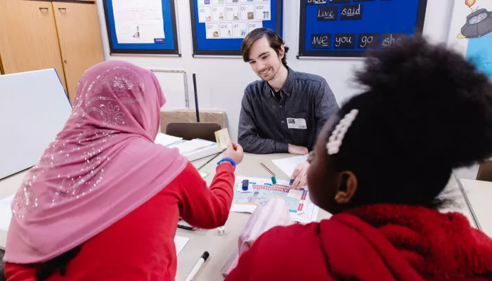 A male tutor, wearing a grey shirt, smiles as two pupils participate in a session.