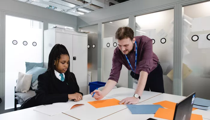 A female secondary school pupil in a dark school uniform is sat at a desk. To her right, a young male tutor wearing a purple shirt is standing over the desk and going through an example on a sheet of paper.