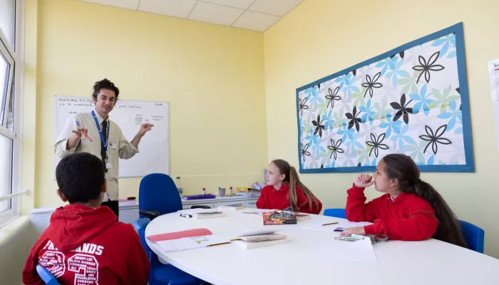 A tutor stands in front of a group of three pupils. The pupils are wearing red jumpers.