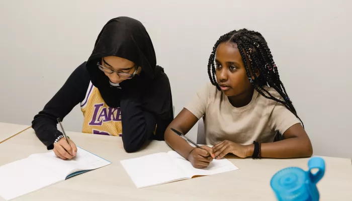 Two secondary-school age female pupils are sat at a desk. The one on the left is wearing a long black top, LA Lakers t-shirt and black hijab, the one on the right has long black braided hair and is wearing a beige t-shirt. They are writing in exercise books.