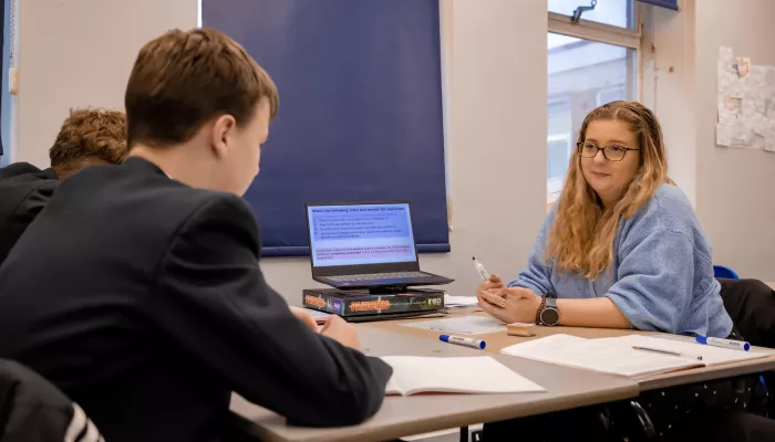 To the left of the photo is a male secondary school-aged pupil wearing a dark blazer. He is turned away from the camera and sat at a desk. To the right is a female tutor with long blonde hair wearing a blue sweater. On the middle of the desk is an open laptop.