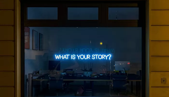 A full-width shop window. In the window is a small blue neon sign that says 'what is your story?' in capital letters.