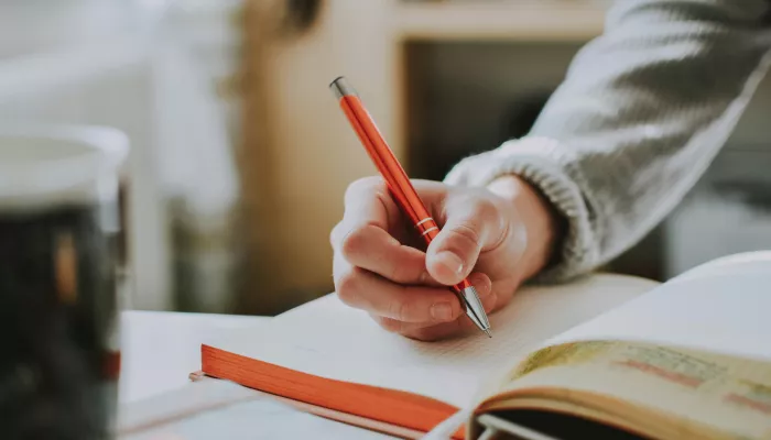 A close up shot of a person's hand above a notebook. The person is wearing a light grey jumper and holding a dark orange pen.