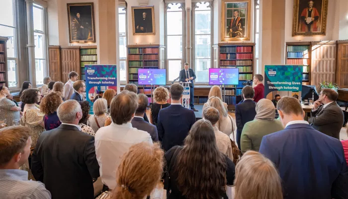 A group of people are stood in a formal room at a Tutor Trust Summer Reception. They are watching a speech from Tutor Trust's CEO, Ed Marsh.