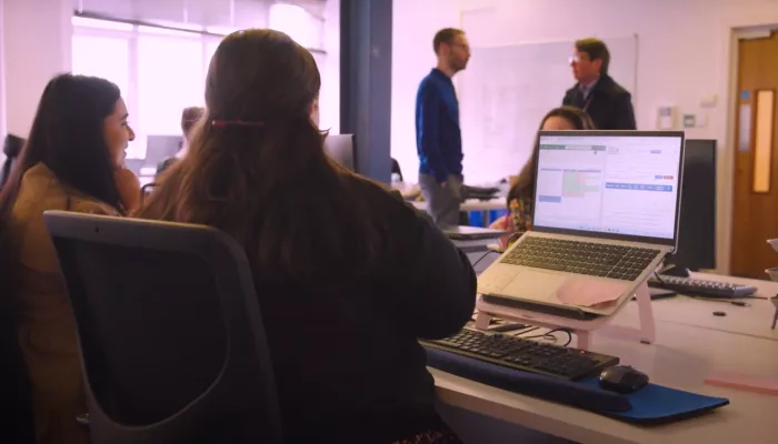 The back of a woman at a desk in an office and two men standing further back talking