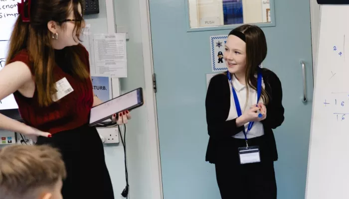 A girl in a school uniform stands in front of a whiteboard, smiling at her tutor who is holding a tablet