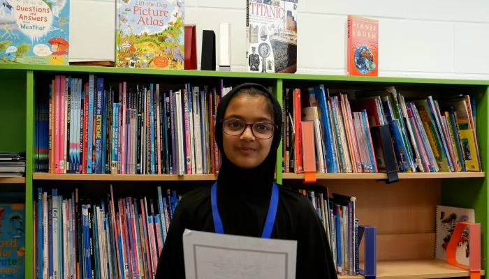 Umme is a primary school pupil and winner of the Speak Up North competition. She wears glasses and smiles to camera while holding and reading her poem.