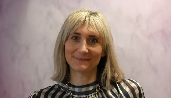 A blonde woman in a checked blouse stands in front of a lilac background