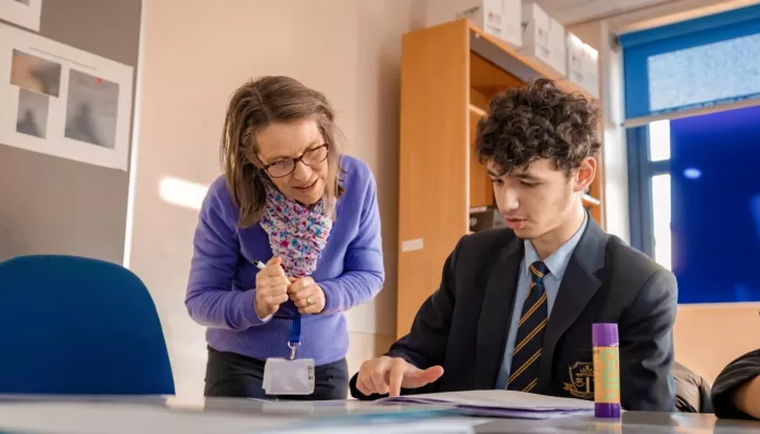 A boy at a table in a school uniform is helped by his female tutor who is standing next to him, wearing a blue jumper