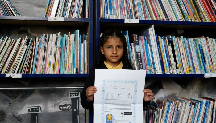 Aisha stands in front of library books and proudly holds up a large, printed version of her winning poem.