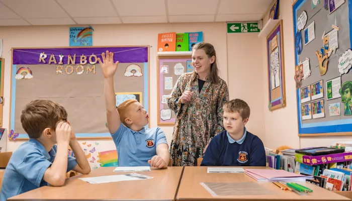 Two children are at a desk, one has his hand up and a teacher stands behind them