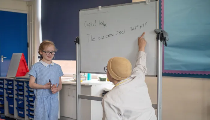 A girl in a blue school uniform and glasses stands in front of a whiteboard looking at her tutor who is wearing a hat and pointing at the whiteboard