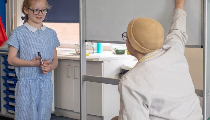 A girl in a blue school uniform and glasses stands in front of a whiteboard looking at her tutor who is wearing a hat and pointing at the whiteboard