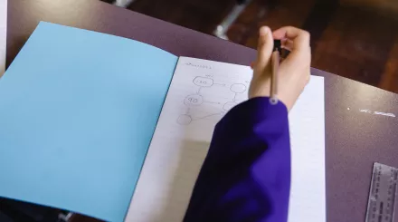 An open workbook on a desk with mathematics work in it. A pupil's arm, wearing a purple blazer, and hand are visible above the book, about to write in it.