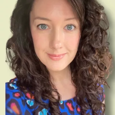 Helen has long dark hair with curls and a floral dress. She looks into the camera smiling.