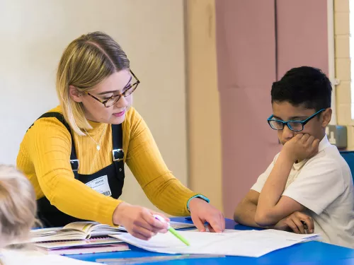 A tutor in a yellow jumper wearing dungarees is explaining a document to a young boy wearing blue glasses.