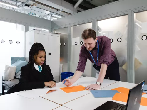 A female secondary school pupil in a dark school uniform is sat at a desk. To her right, a young male tutor wearing a purple shirt is standing over the desk and going through an example on a sheet of paper. 