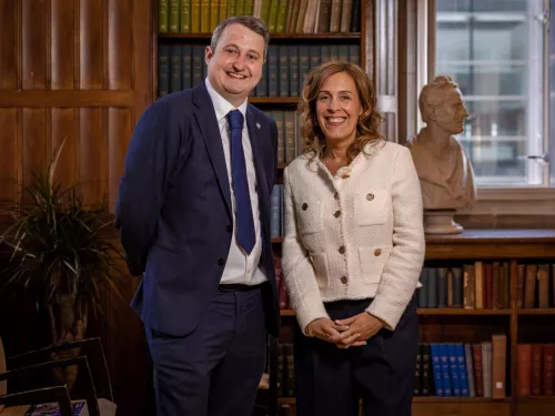 Ed in a dark blue suit and blue tie, stands next to Abigail, in a white jacket and dark trousers. They are both standing in front of a bookcase and both are smiling for the camera.