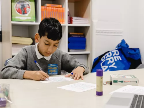 A primary school-aged boy wearing a grey school jumper is sat at a desk. He is writing with a pen on a sheet of paper and concentrating hard.