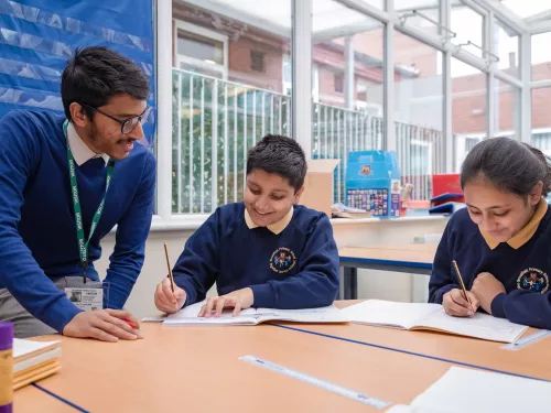 A male tutor with black hair and glasses, wearing a blue jumper, is leaning over a desk. Two primary aged pupils, a boy and a girl, wearing dark blue school jumpers are holding pencils and writing in exercise books. Everyone is smiling.