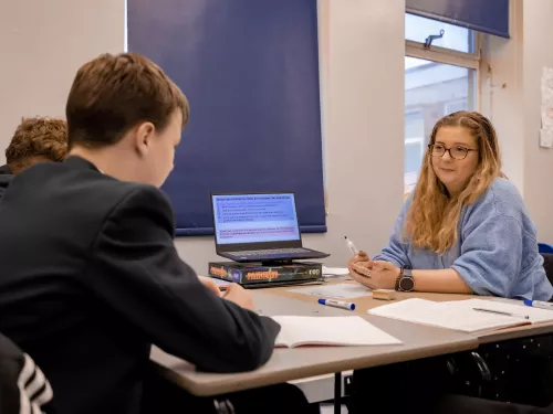 A male secondary school pupil wearing a dark blue school blazer is sat at a desk. On the other side of the desk is a female tutor with long blonde hair and glasses wearing a pale blue jumper. A laptop computer is on the middle of the desk. 