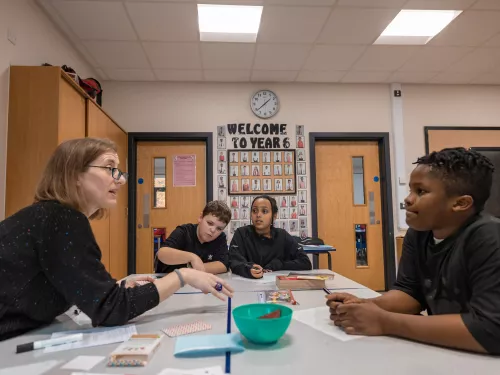 A female tutor wearing glasses is sat with three primary aged pupils around a desk. She is explaining a concept. The pupils are listening. In the back there is a colourful 'Welcome to Year 6' display on the wall.