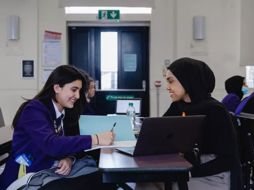 A secondary aged female pupil with long dark hair, wearing a purple blazer, is sat at a desk writing. An open laptop is on the desk. To the right of the desk is sat a young female tutor wearing a black jumper and black hijab.