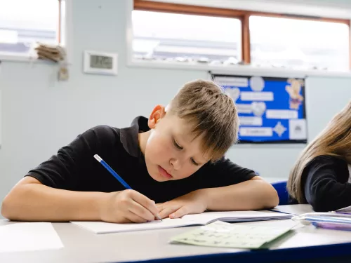 A primary school-aged boy wearing a black t-shirt is sat at a desk and is writing with a pencil in an exercise book.