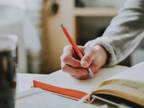 A close up shot of a person's hand above a notebook. The person is wearing a light grey jumper and holding a dark orange pen.