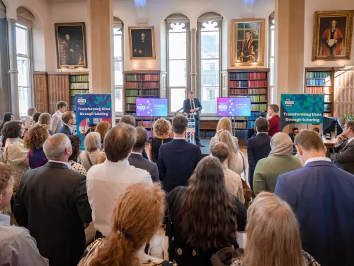 A group of people are stood in a formal room at a Tutor Trust Summer Reception. They are watching a speech from Tutor Trust's CEO, Ed Marsh.