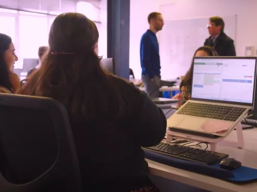 The back of a woman at a desk in an office and two men standing further back talking