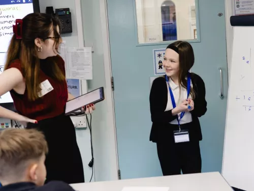 A girl in a school uniform stands in front of a whiteboard, smiling at her tutor who is holding a tablet