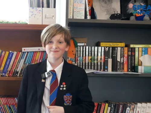 Pupil James wears a black blazer with several achievement badges on the sides, white shirt, and blue and red striped tie. He excitedly tells us how it feels to be a competition winner.