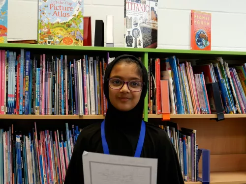 Umme is a primary school pupil and winner of the Speak Up North competition. She wears glasses and smiles to camera while holding and reading her poem.