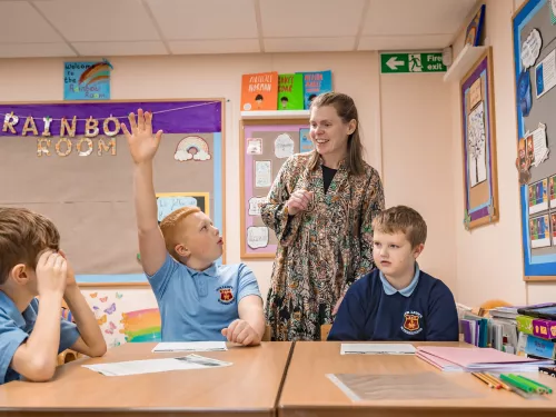 Two children are at a desk, one has his hand up and a teacher stands behind them