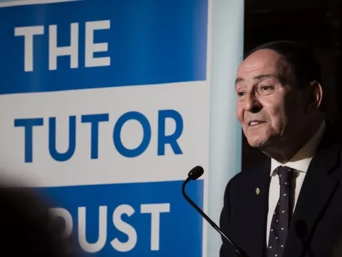 A man stands behind a microphone and next to a large Tutor Trust logo