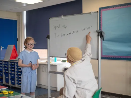 A girl in a blue school uniform and glasses stands in front of a whiteboard looking at her tutor who is wearing a hat and pointing at the whiteboard