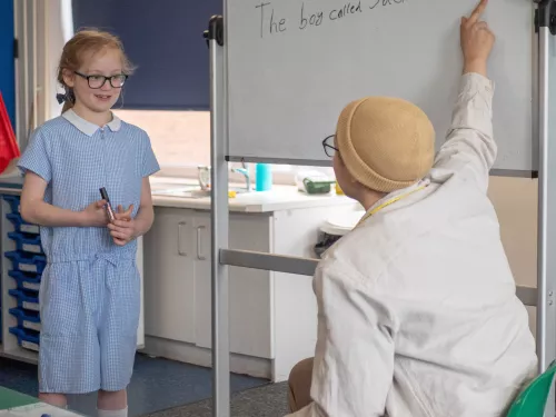 A girl in a blue school uniform and glasses stands in front of a whiteboard looking at her tutor who is wearing a hat and pointing at the whiteboard